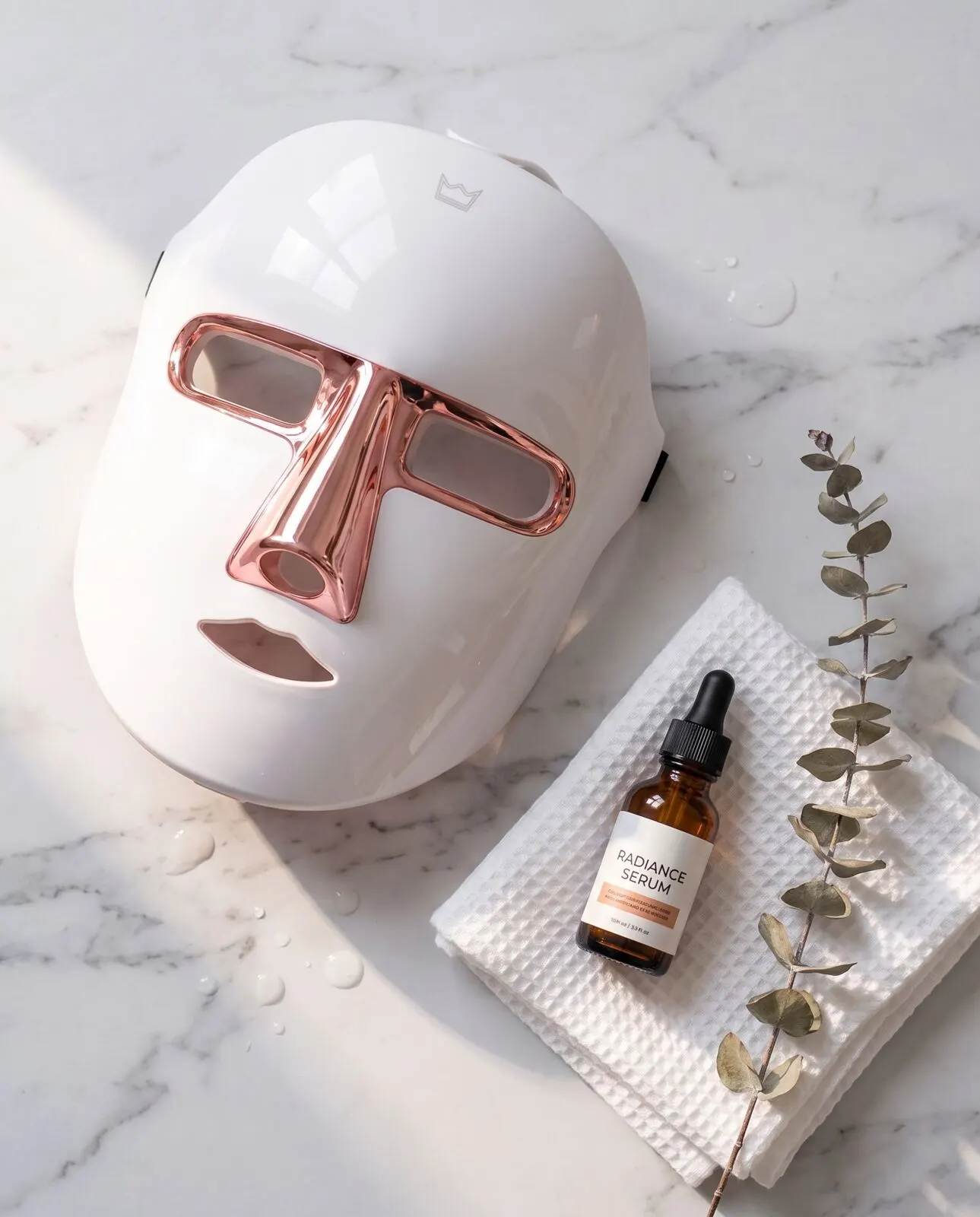 Lumoras LED face mask laid on marble beside a serum bottle, a waffle towel and eucalyptus